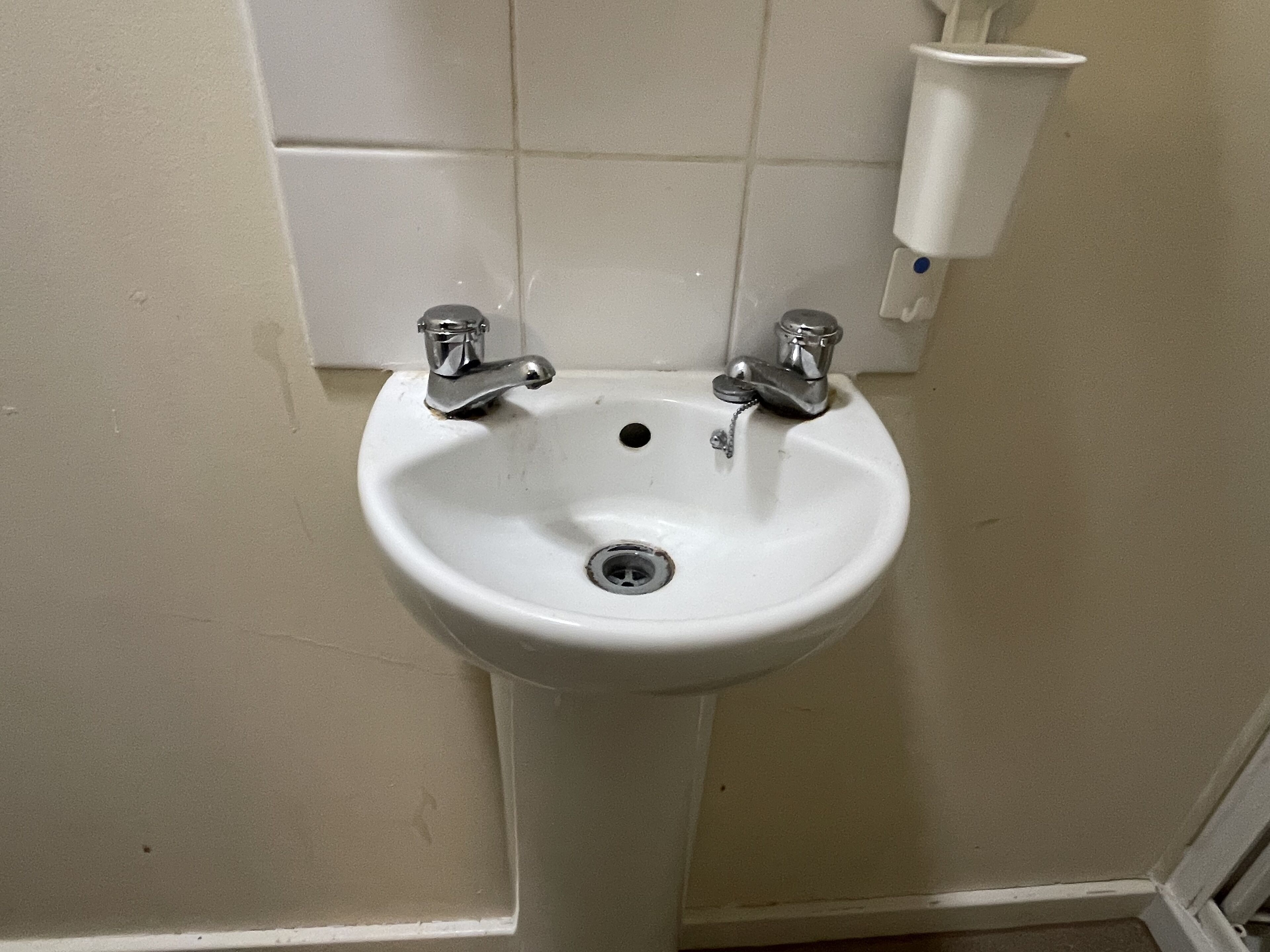 Uma pia de pedestal branca com duas torneiras de metal cromado, um ralo e um dosador de sabonete fixado na parede.