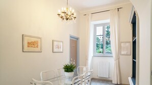 Un coin repas avec une table en verre et des chaises blanches, un lustre, des tableaux encadrés au mur et une fenêtre avec des rideaux.