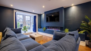 A modern living room with a dark blue accent wall, a flat-screen TV mounted above a fireplace, and a large window with blue curtains. - Modern 6BR/4Bath in the heart of #LakeDistrict (Cumbria)