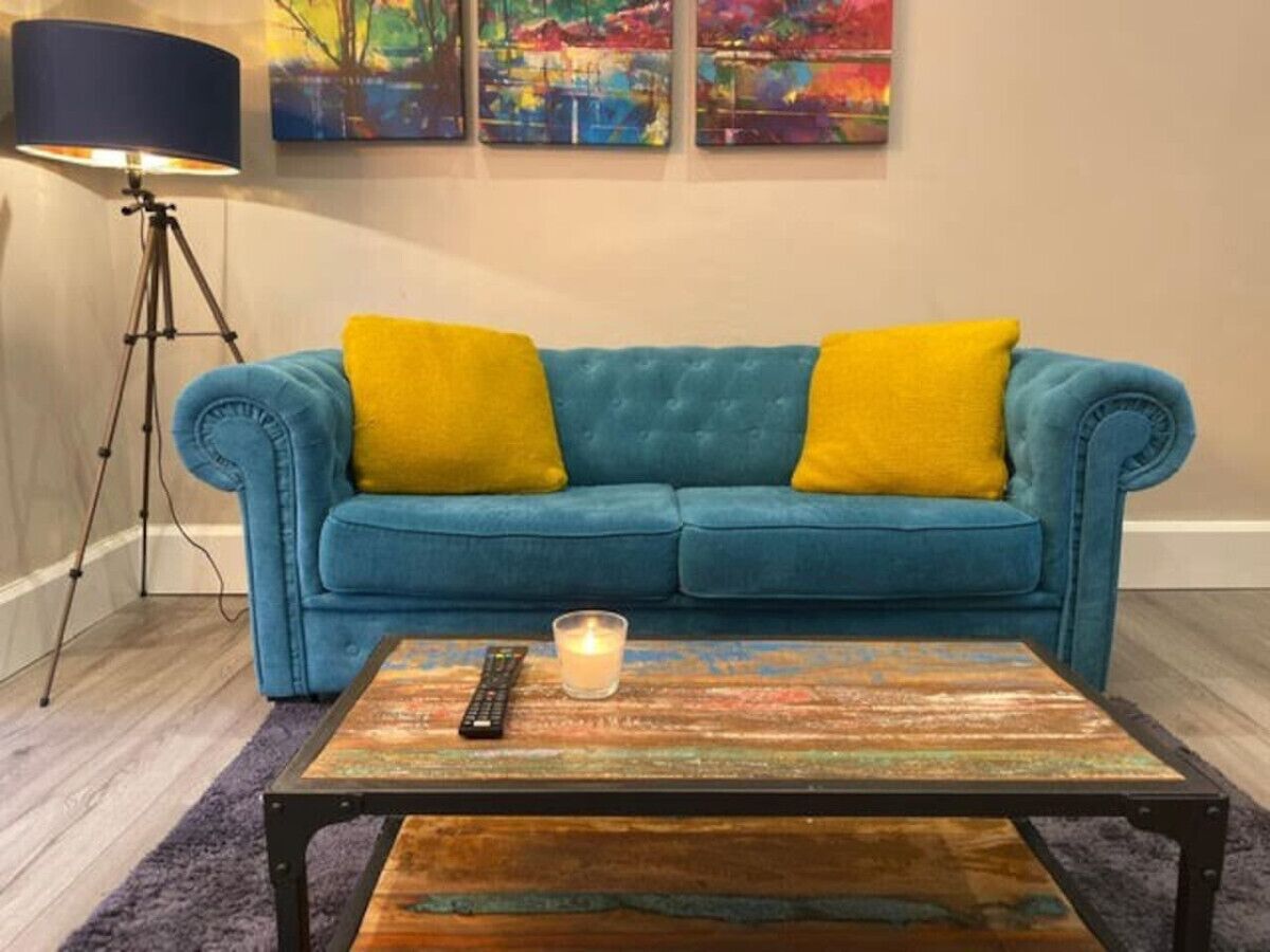 Ein blaues Sofa mit gelben Kissen, ein Couchtisch aus Holz mit Fernbedienung und Kerze sowie eine Stehlampe.