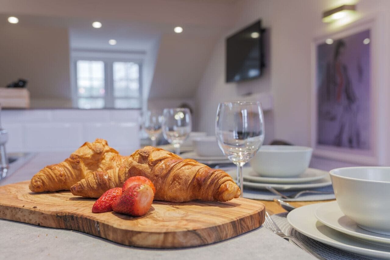 Un desayuno con cruasanes y una fresa sobre una tabla de madera, un vaso de agua, y platos y tazones blancos en una mesa.