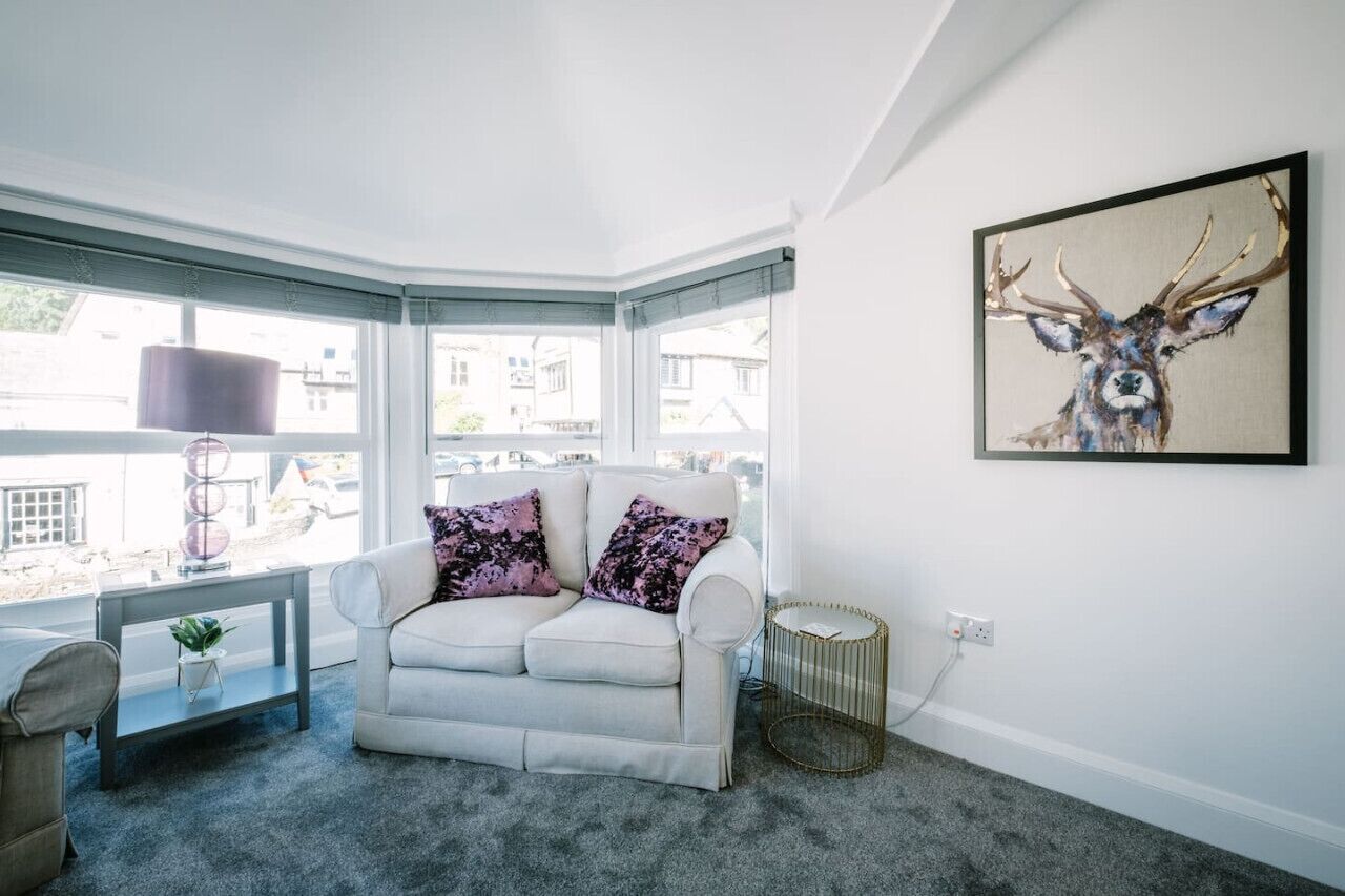 A corner room with a white sofa, a small table with a lamp, a round side table, and a large window.