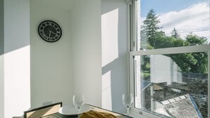 Ein Essbereich mit Tisch für zwei Personen, einer Wanduhr und Blick auf Bäume durch ein Fenster.