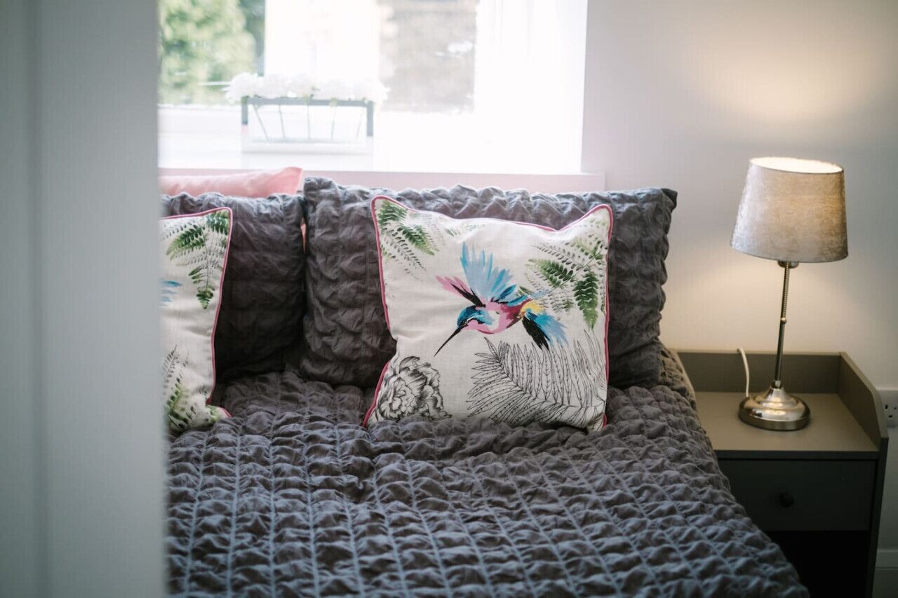 Uma cama com um cobertor cinza texturizado, duas almofadas com desenho de pássaros, uma luminária de cabeceira e uma janela com vista para vegetação.