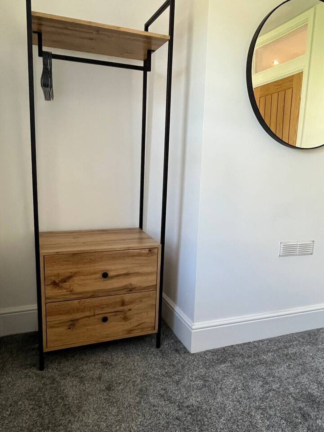 A room with a wooden bench with drawers, a round mirror on the wall, and a vent near the floor.