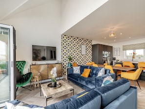 A modern living room with a sofa, coffee table, and a TV. There is a dining area with yellow chairs and a kitchen in the background.