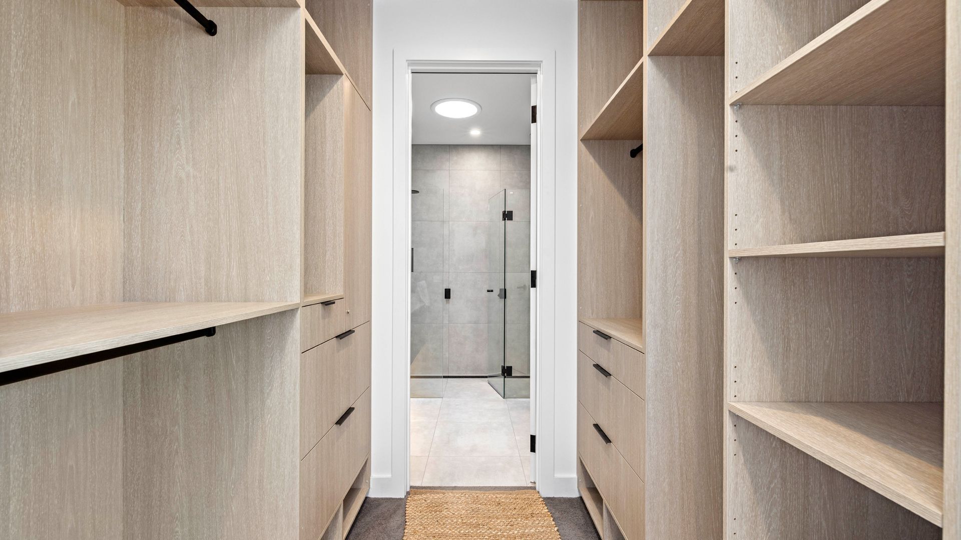 A narrow hallway with wooden shelving units on both sides, leading to a room with a tiled floor and a door with a glass panel.