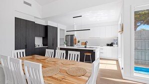 A modern kitchen with white cabinets, a stainless steel refrigerator, and a wooden dining table with white chairs. - The Sanctuary - Rosebud (Rosebud)