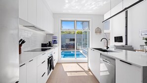 A modern kitchen with white cabinetry, a built-in dishwasher, and a large window overlooking a pool. - The Sanctuary - Rosebud (Rosebud)