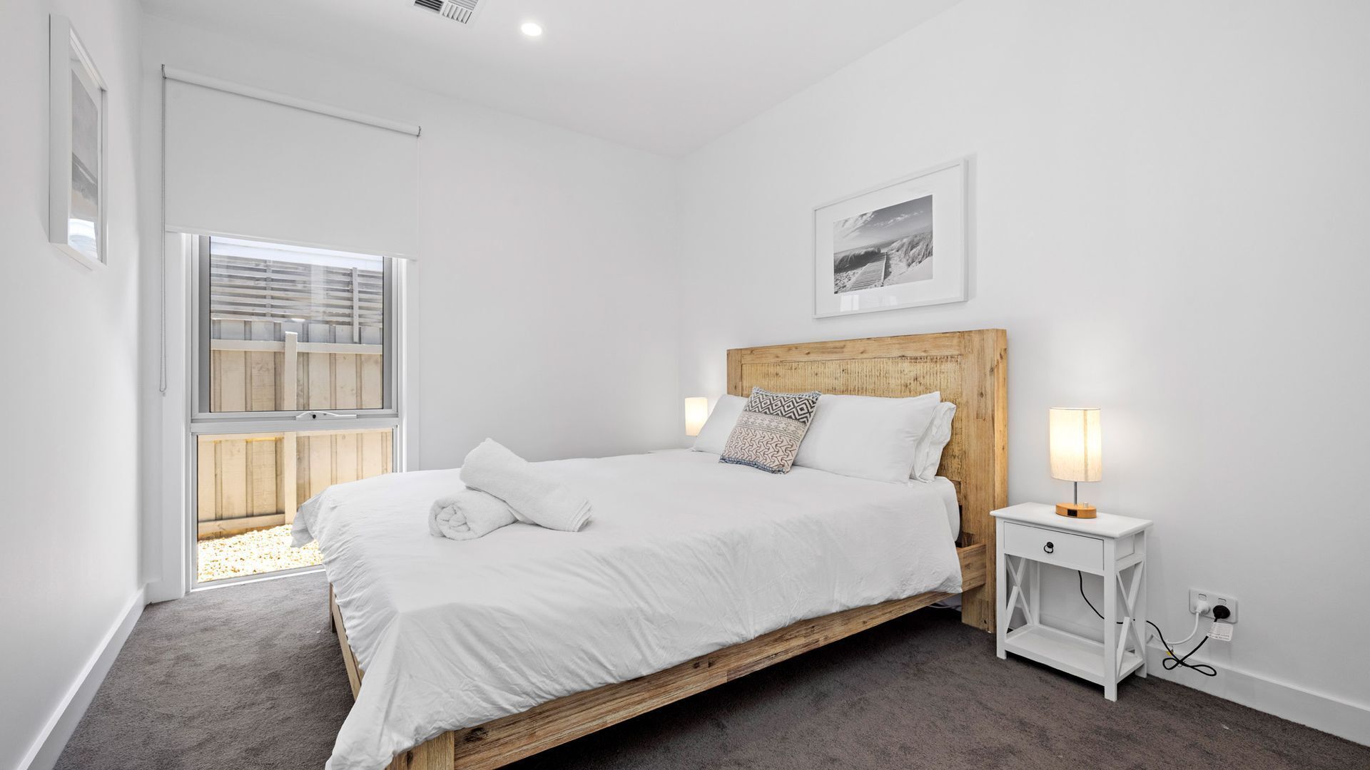 Ein Schlafzimmer mit einem hölzernen Bett, weißer Bettwäsche, einem Nachttisch mit Lampe, einem Fenster mit Jalousien und einem gerahmten Bild an der Wand.
