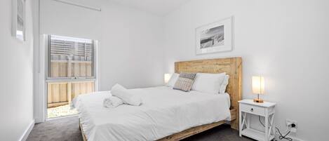 Ein Schlafzimmer mit einem hölzernen Bett, weißer Bettwäsche, einem Nachttisch mit Lampe, einem Fenster mit Jalousien und einem gerahmten Bild an der Wand.
