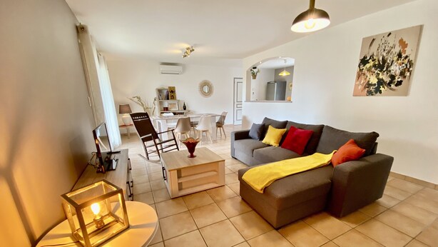 A modern living room with a grey sofa, a small round table with a lamp, and a dining area in the background. - Villa 6p, 3ch Avec Grand Jardin Arboré Clos, Clim, Village Classé de Vogue (Vogüé)