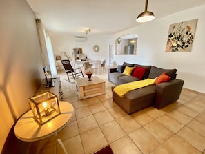 A modern living room with a grey sofa, a small round table with a lamp, and a dining area in the background.