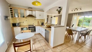 A modern kitchen with white cabinets, a central island, a dining area with a table and chairs, and a view of the outdoors through a large window. - Villa 6p, 3ch Avec Grand Jardin Arboré Clos, Clim, Village Classé de Vogue (Vogüé)
