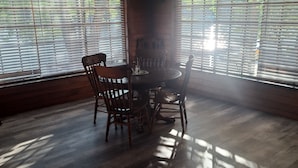 Un comedor con una mesa redonda y cuatro sillas, suelo de madera y ventanas amplias con persianas.