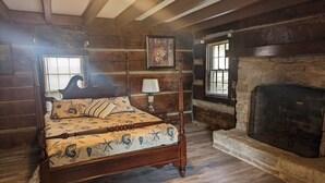 Una cama con dosel con ropa de cama floral, una mesita de noche de madera, una lámpara y una chimenea de piedra en una habitación rústica con paredes y pisos de madera.