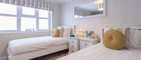 A hotel room with two single beds, a nightstand with a lamp, a mirror, and a window with curtains.