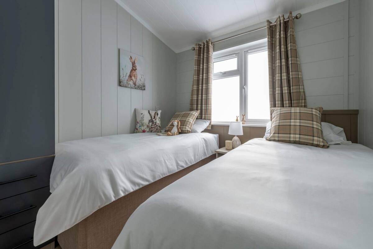 A bedroom with two beds, a window with curtains, a bedside table, and a wall-mounted picture.