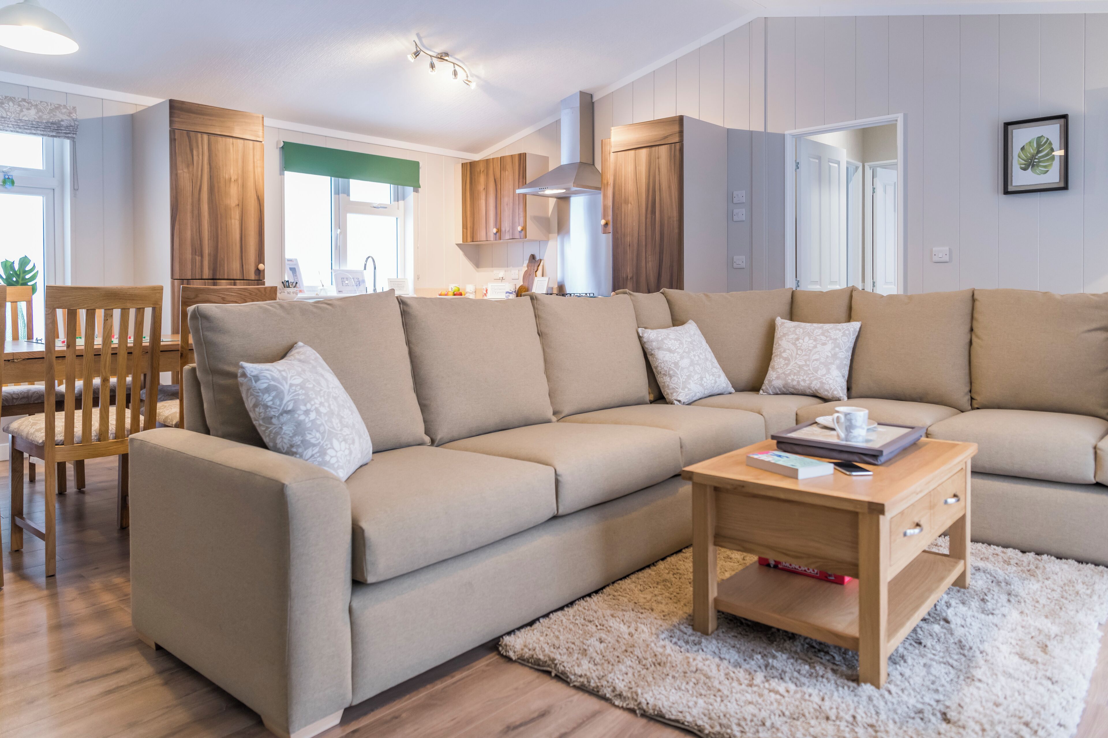 A modern living room with a beige sectional sofa, wooden cabinets, a dining area with a wooden table and chairs, and a kitchen in the background.