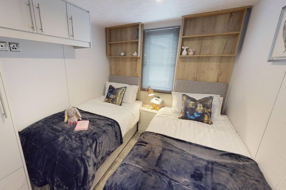 A bedroom with two beds, a wardrobe, a shelf with decorative items, and a window with blinds.