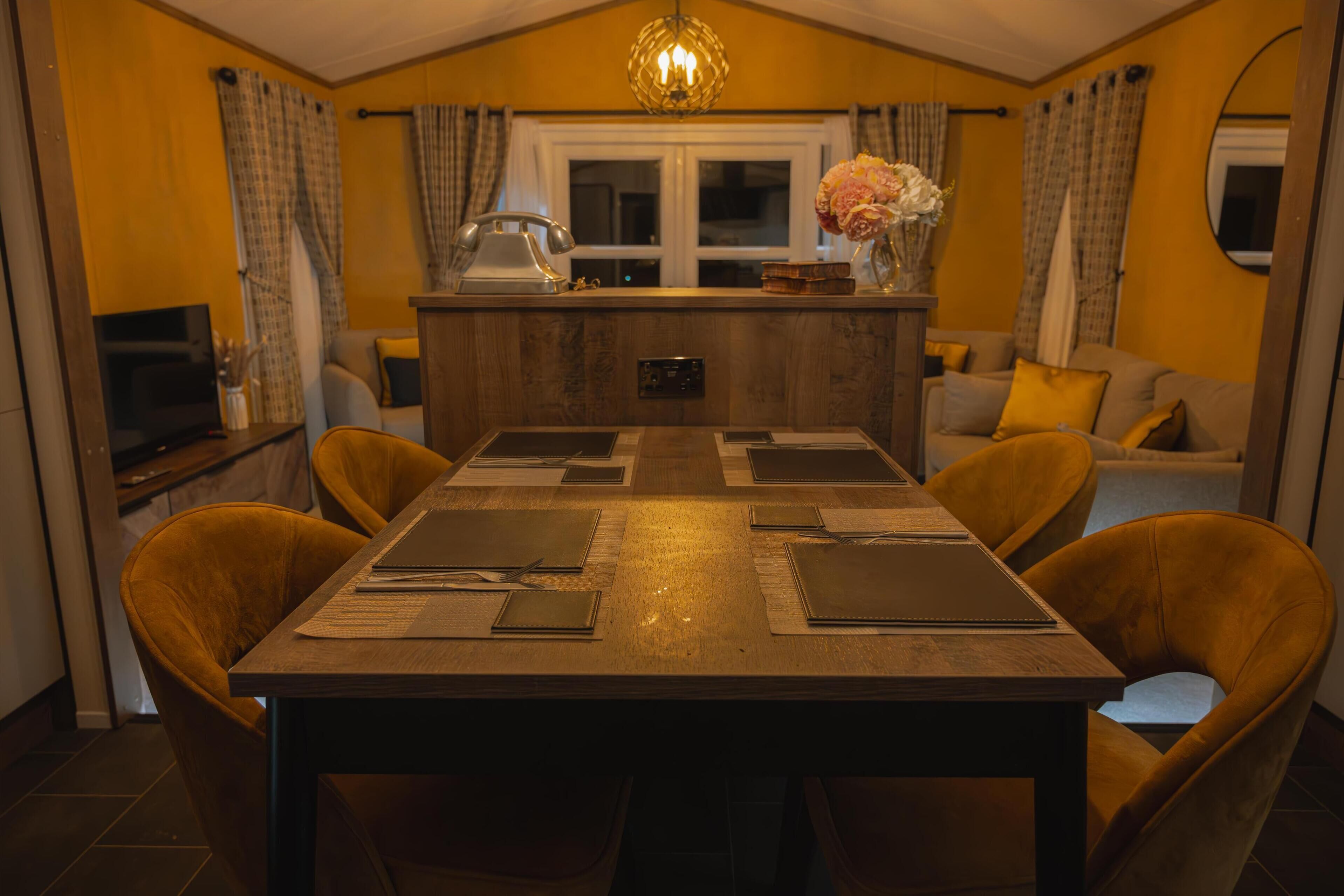 A dining area with a wooden table set for four, surrounded by yellow chairs. A chandelier hangs from the ceiling, and a kitchen area is visible in the background.
