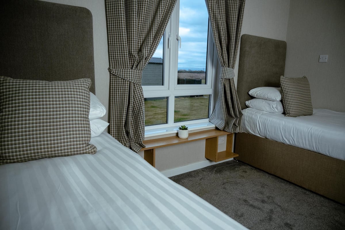A bedroom with two beds, a window with curtains, a small plant on the windowsill, and a view of the outdoors.