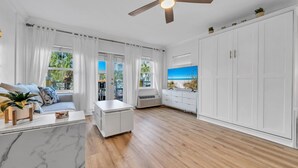 A spacious living room with a wooden floor, white furniture, a ceiling fan, and a large window with a view of the outdoors.