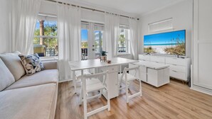 A bright, white room with a wooden floor, a white sofa, a small table, and a large TV screen displaying a beach scene.