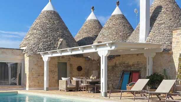 Un espace aménagé au bord de la piscine, avec un bâtiment en pierre doté de toits coniques, un espace salon couvert et des chaises longues.