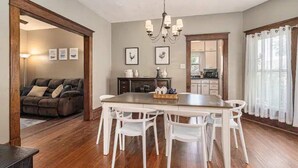 A dining area with a wooden table and white chairs, a chandelier, and framed artwork on the walls. - Renovated Victorian Home with Private Garden & Fire Pit! (Three Oaks)