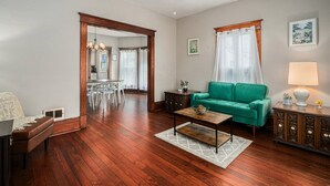 A spacious living room with hardwood flooring, a green sofa, a coffee table, and a dining area with a chandelier. - Renovated Victorian Home with Private Garden & Fire Pit! (Three Oaks)