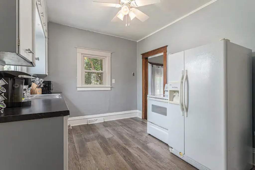 A kitchen with grey walls, a window, a ceiling fan, and a white refrigerator.
