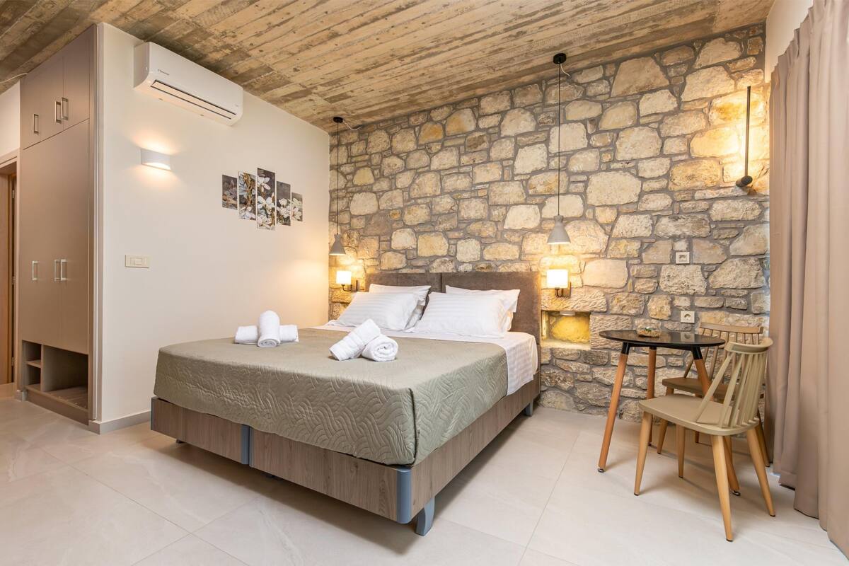 A bedroom with a stone wall, a bed with a wooden headboard, a small table with a chair, and an air conditioning unit.