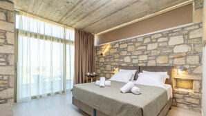 A bedroom with a stone wall, a large bed with white linens, and a view of the outdoors through a sliding door. - Achatis Apartments with sea view (Platanias)