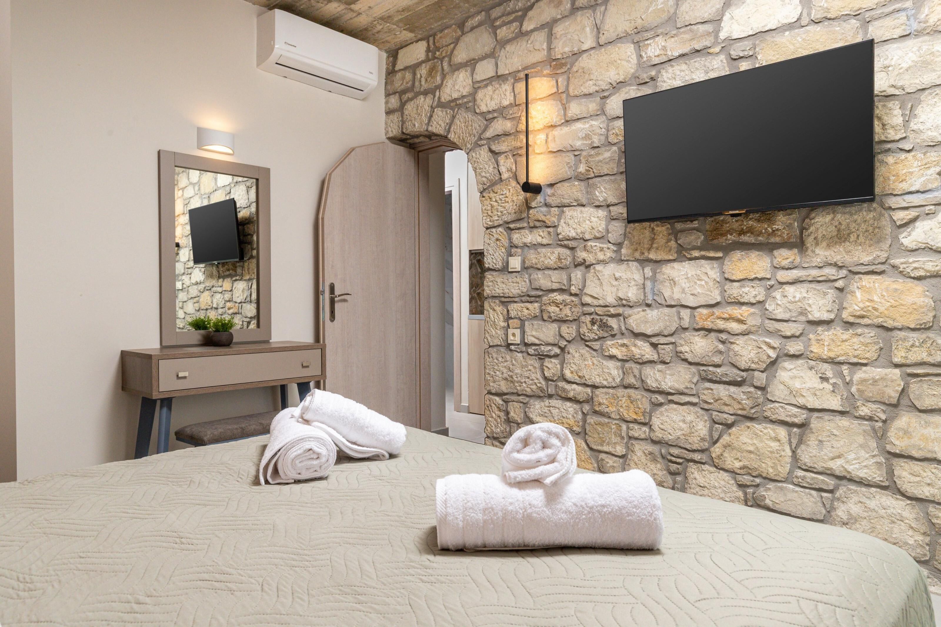 A hotel room with a stone wall, a flat-screen TV mounted above a wooden wardrobe, a bed with a rolled white towel, and a small potted plant on the dresser.