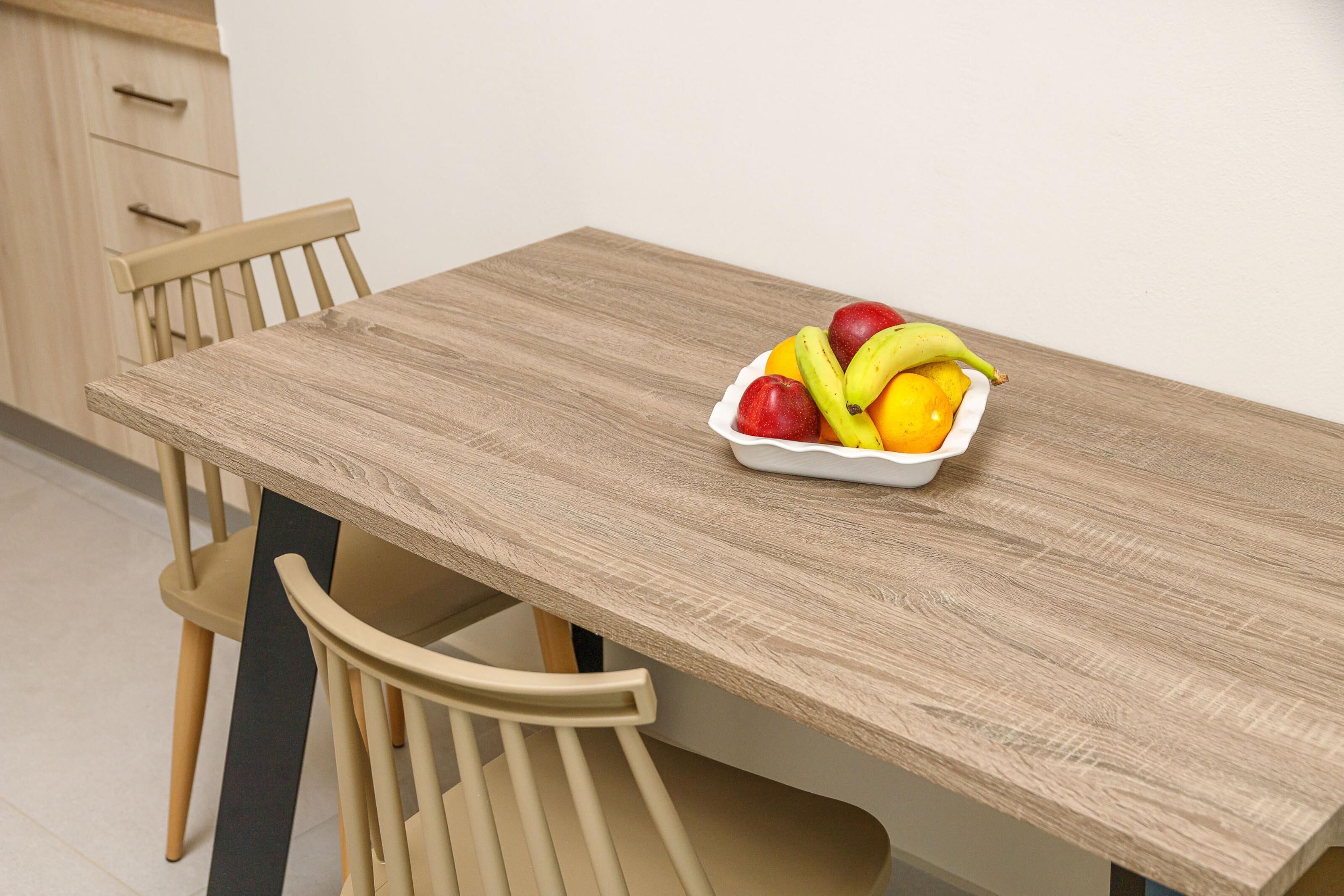 A wooden dining table with a fruit bowl containing apples, bananas, and oranges.