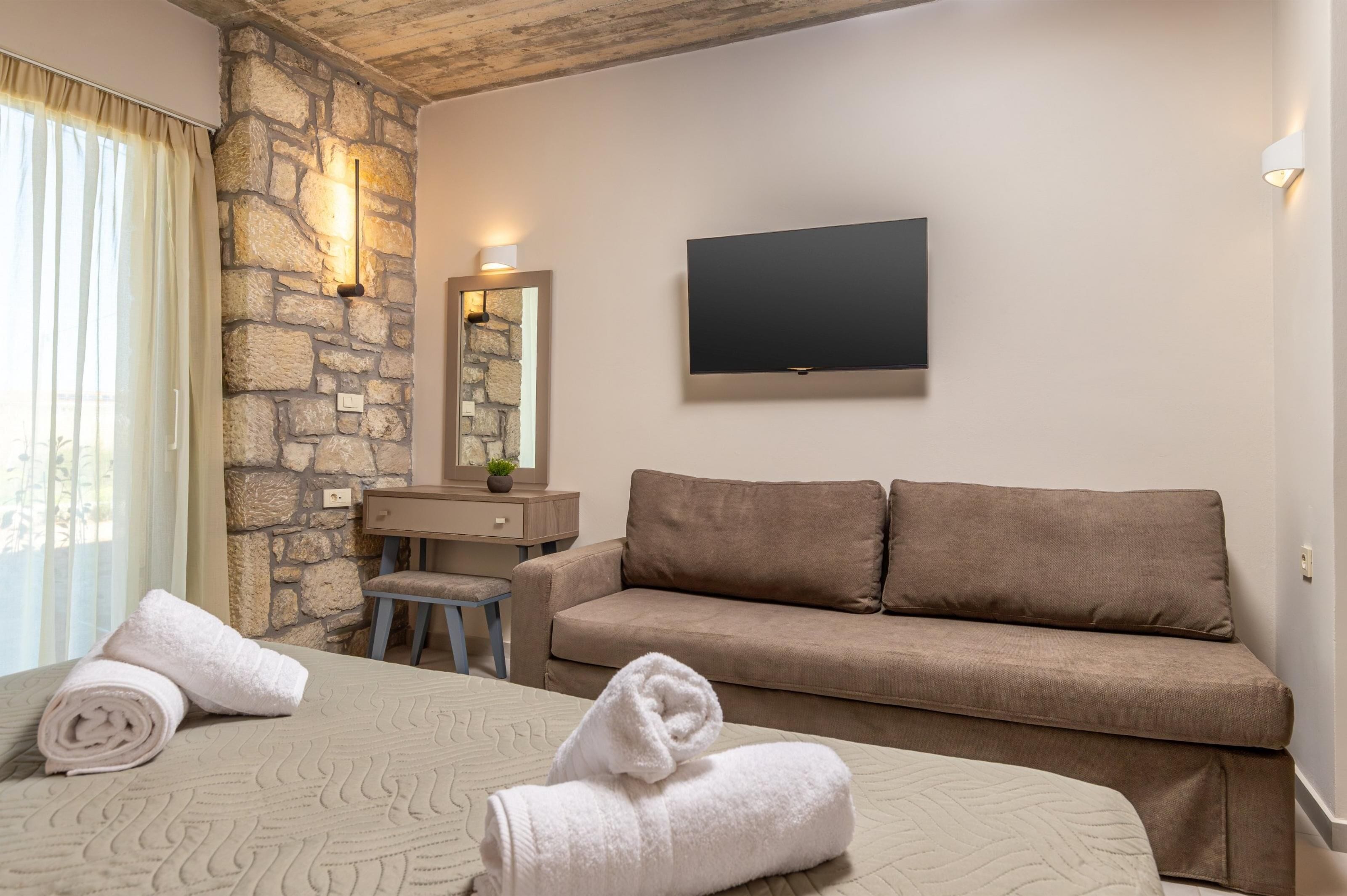 A hotel room with a stone wall, a flat-screen TV, a sofa, a desk, and two rolled towels on the bed.