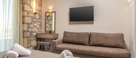 A hotel room with a stone wall, a flat-screen TV, a sofa, a desk, and two rolled towels on the bed.