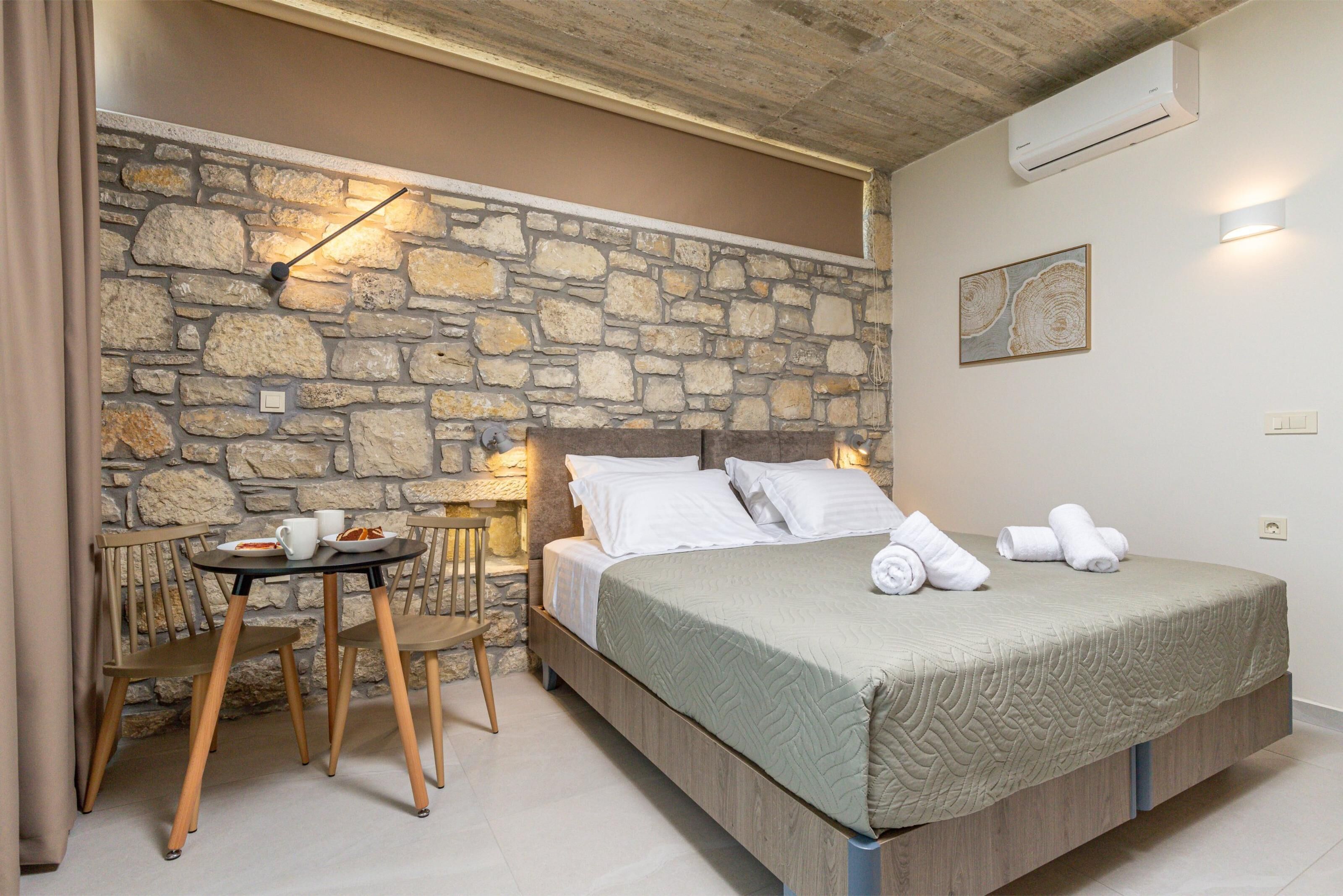 A bedroom with a stone wall, a bed with white linens, a small table with chairs, and a wall-mounted light fixture.