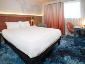 Ein Hotelzimmer mit einem großen Bett, einem Schreibtisch, einem Sessel und Blick ins Freie.