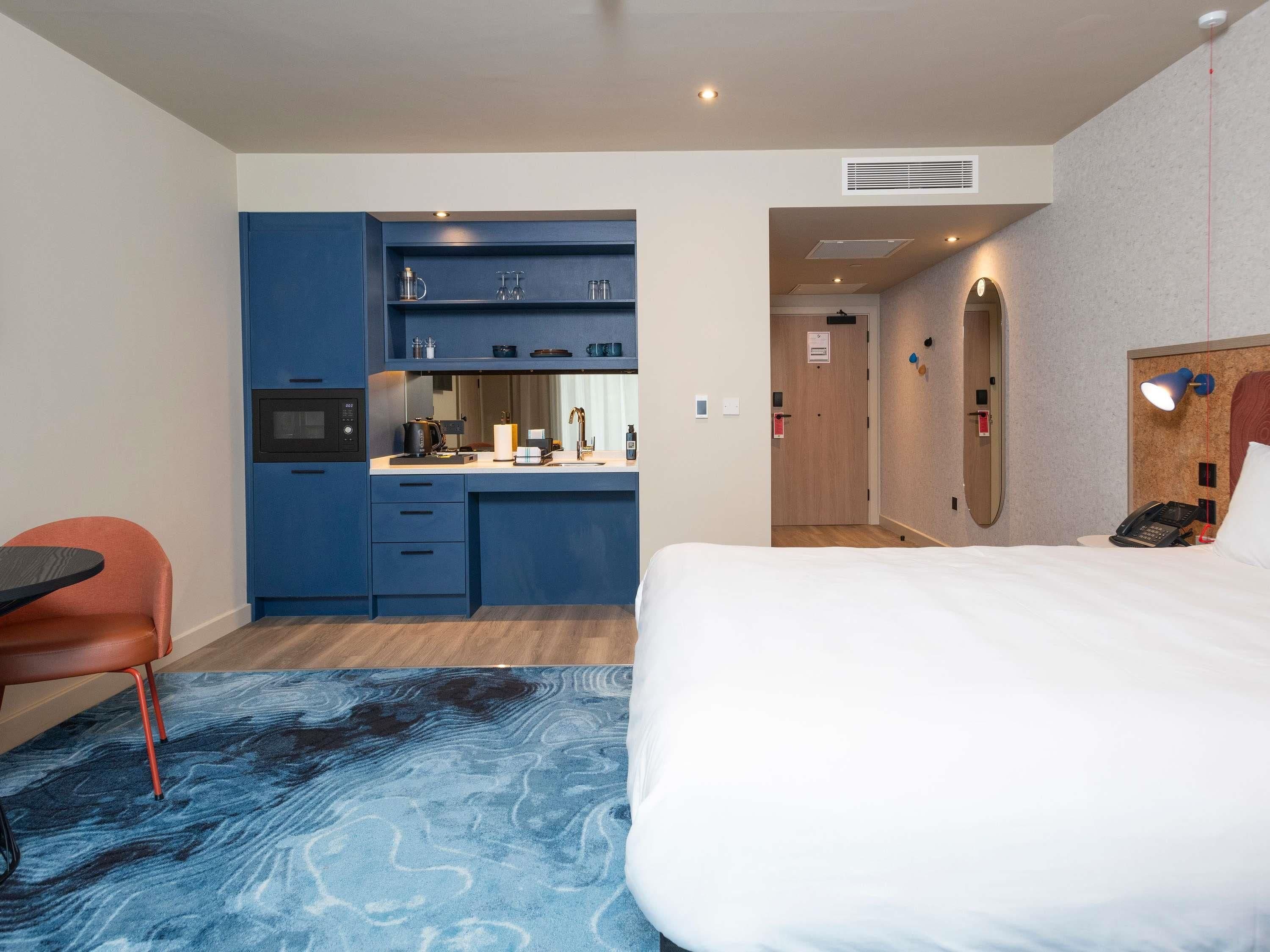Una habitación de hotel con cama, escritorio, silla, kitchenette y baño.