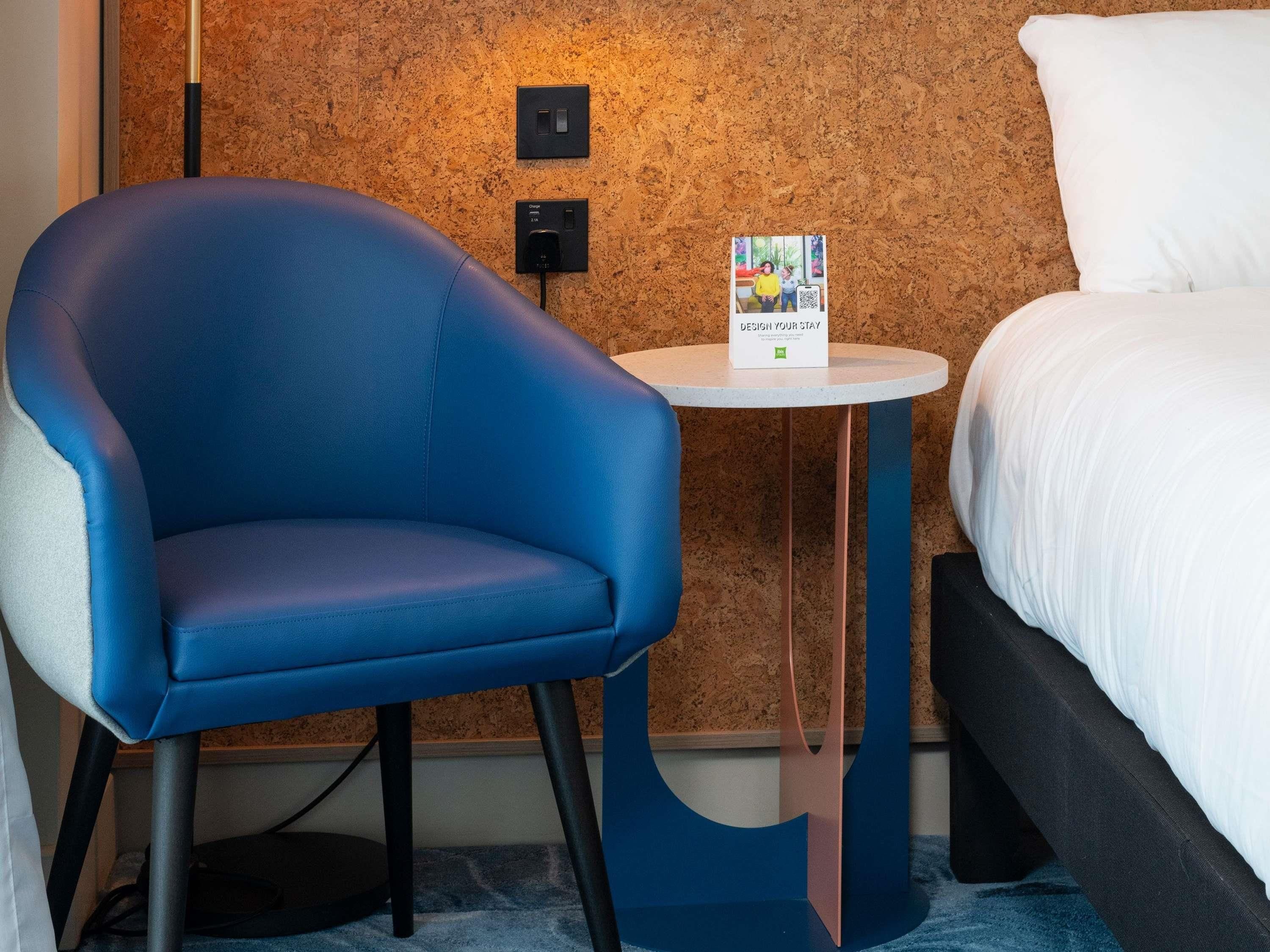 A hotel room with a blue armchair, a small round table with a brochure, and a bed with white bedding.