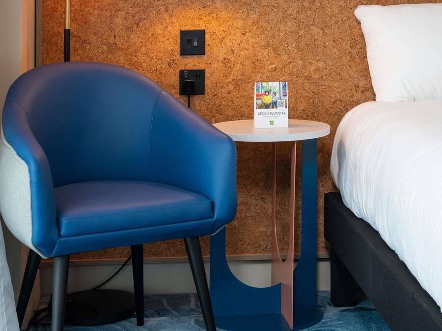 Ein Hotelzimmer mit blauem Sessel, einem kleinen runden Tisch mit einer Broschüre und einem Bett mit weißer Bettwäsche.