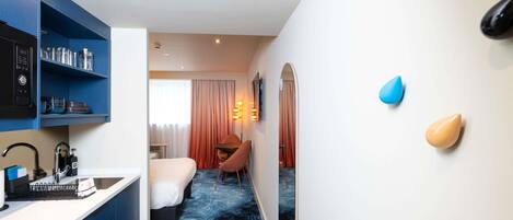 A hotel room with a kitchenette, a bed, a desk, and a bathroom.