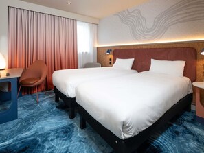 Ein Hotelzimmer mit zwei Betten, einem Schreibtisch, einem Stuhl und einem großen Fenster mit Vorhängen.