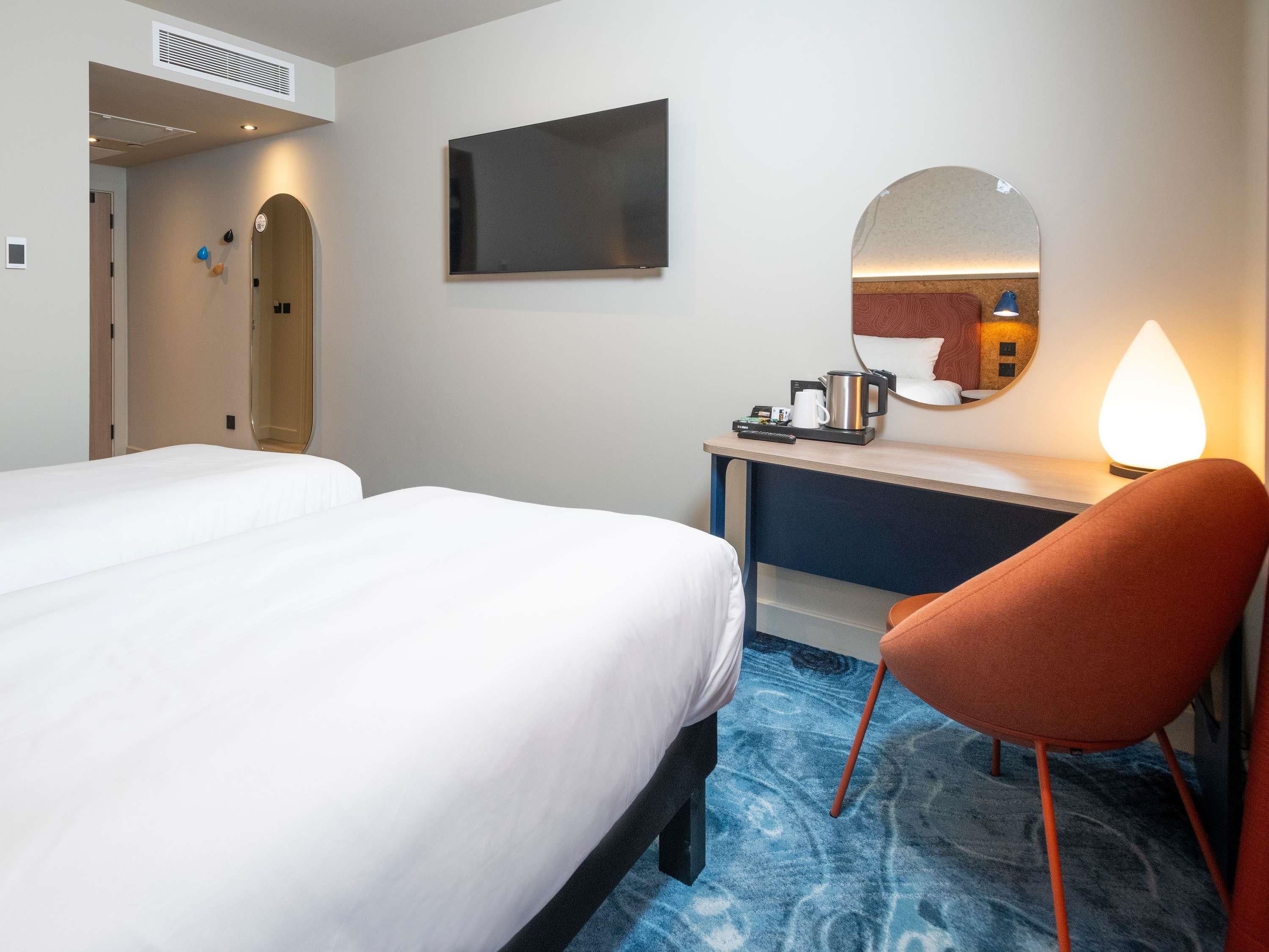 A hotel room with two beds, a desk, a chair, a mirror, and a coffee maker.