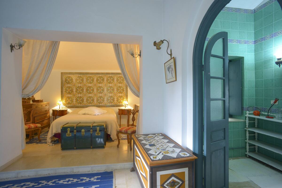 A bedroom with a bed, curtains, a dresser, a chair, and a small table with a lamp.