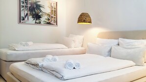 Een hotelkamer met twee bedden, elk voorzien van witte lakens en opgerolde handdoeken, een geweven lamp en een lijst met daarop een afbeelding van palmbomen.