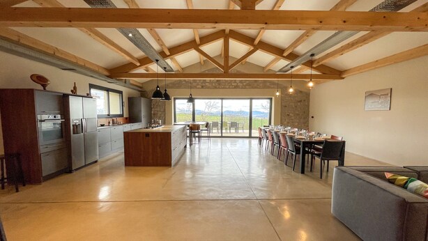 A spacious kitchen with a central island, wooden beams, and a dining area with a view of the outdoors. - Gîte des Ollières : Entre Charme Traditionnel et Confort Moderne (Monistrol-sur-Loire)