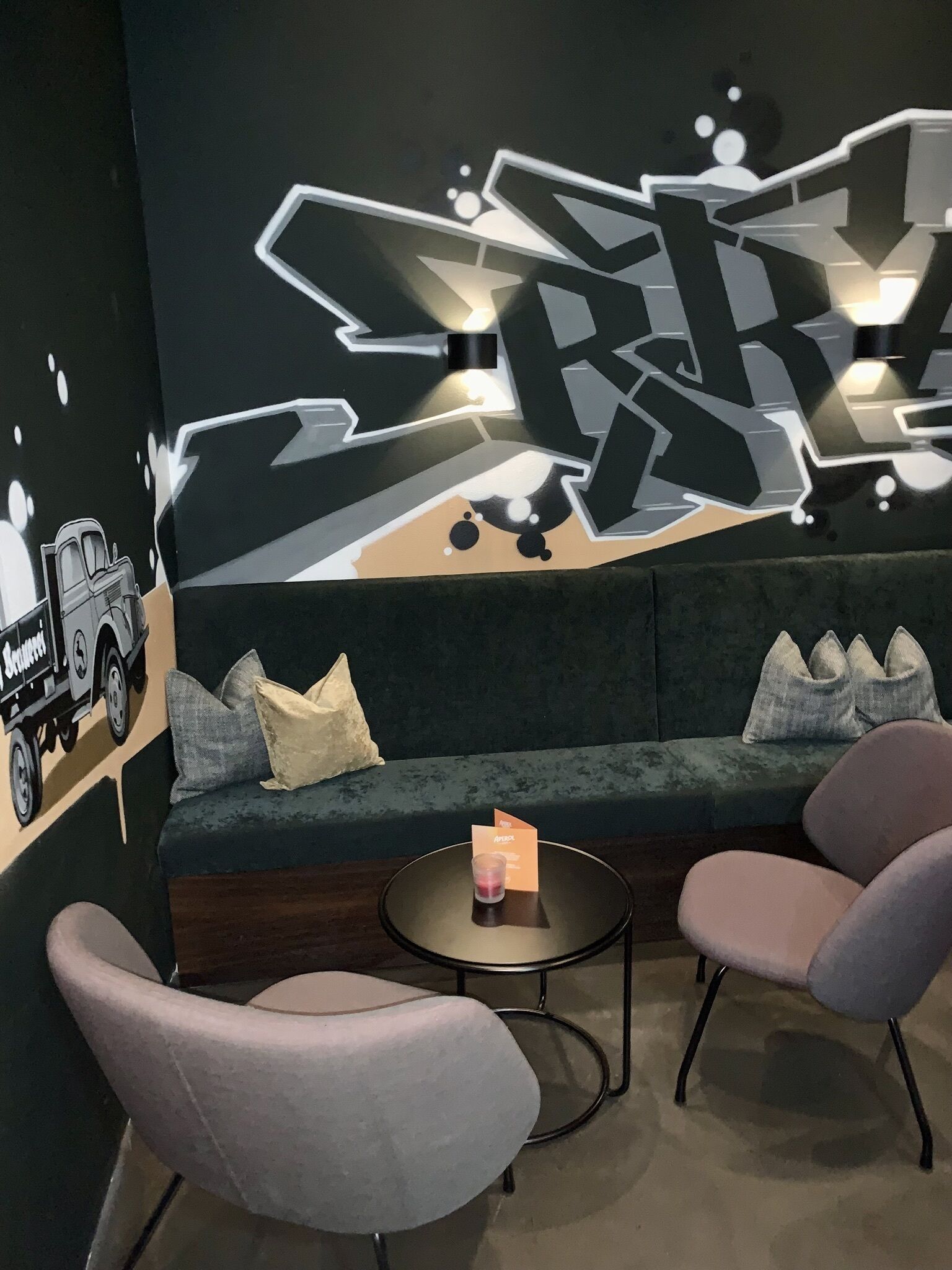 Ein moderner Loungebereich mit einem dunkelgrünen Sofa, zwei Sesseln und einem kleinen runden Tisch. Ein großes abstraktes Wandbild ist zu sehen.